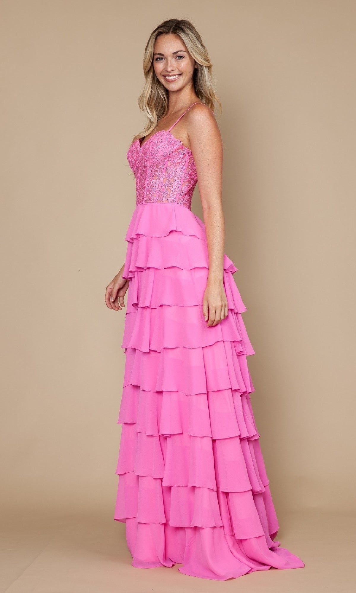 A woman smiles while wearing the Long Tiered Corset Prom Dress: Poly USA 9698, a floor-length pink A-line gown with ruffle tiers and a fitted corset bodice, standing against a plain beige background.